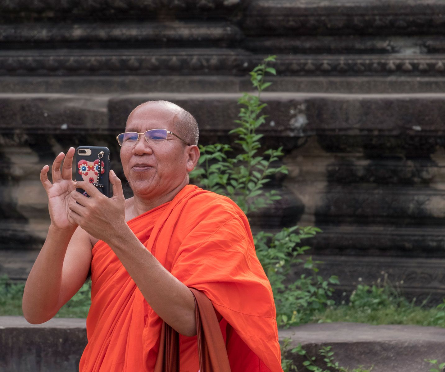 Buddhist monk in orange robes photographing with a phone in front of a stone temple