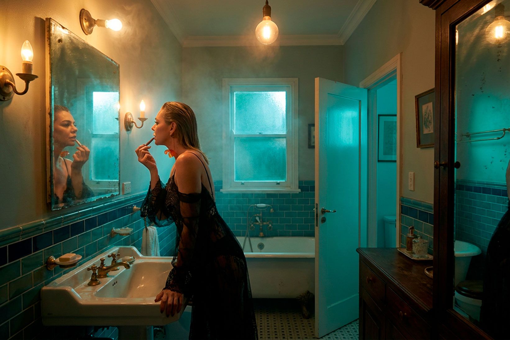 Woman applying lipstick at a bathroom mirror under warm lamps, teal walls — cinematic photography by Claudio Siracusano